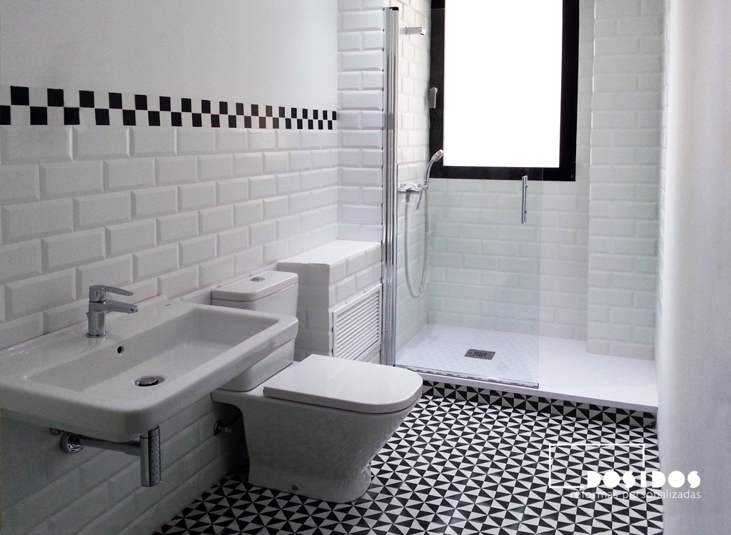 Baño vintage con azulejos blancos biselados y suelo negro con dibujos. Inodoro, lavabo y ducha blancos.